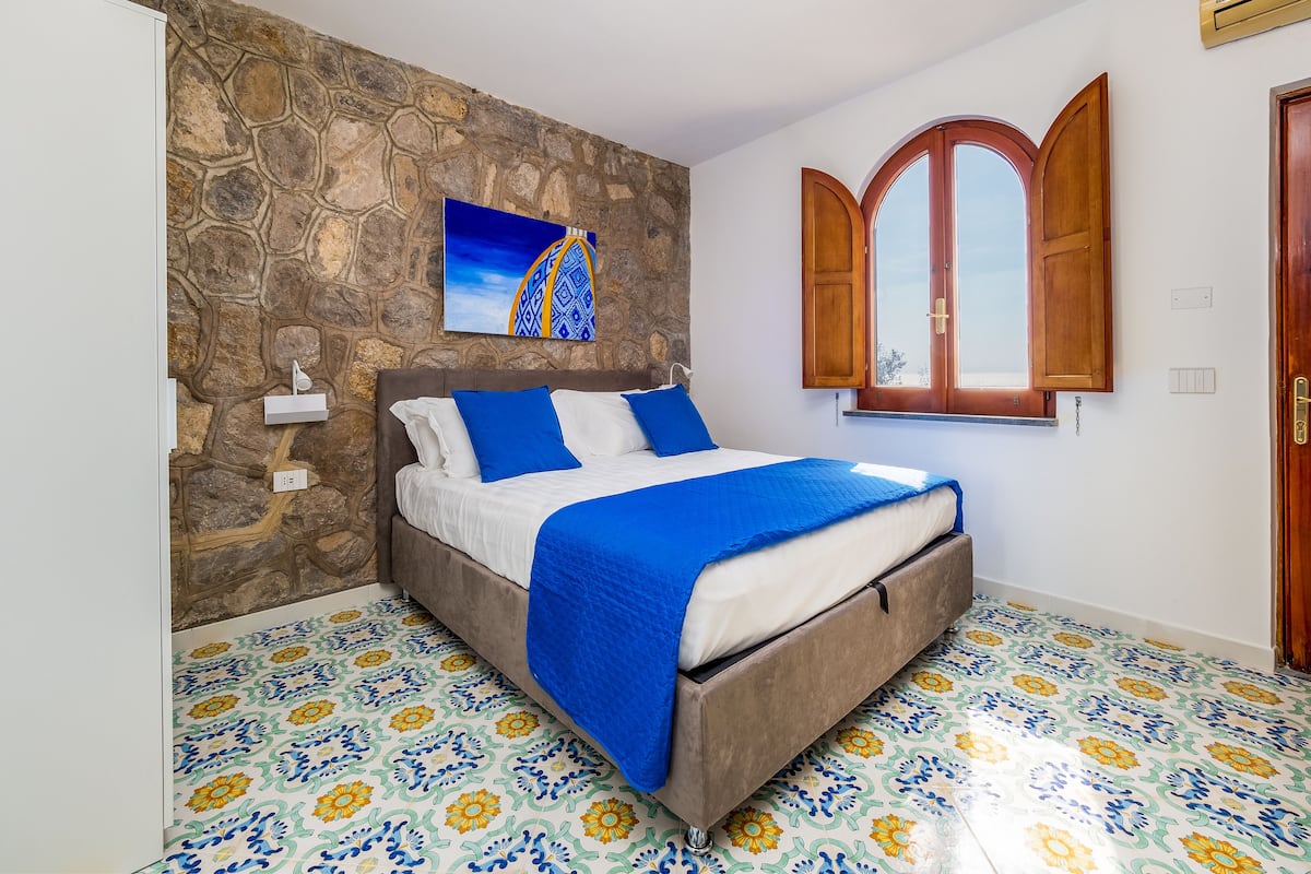 A bedroom with a stone wall, a bed with blue bedding, a window with wooden shutters, and a patterned floor.