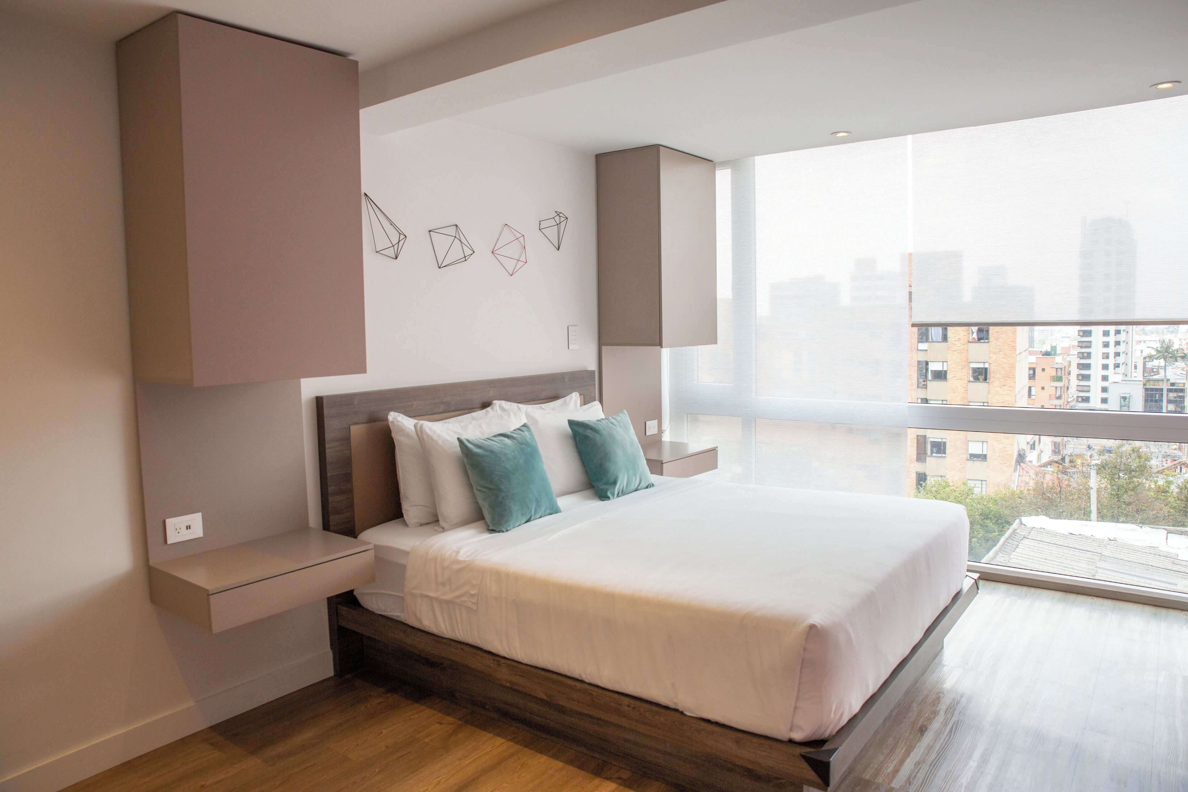 A modern hotel room with a large bed, built-in nightstands, a city view, and geometric wall art.