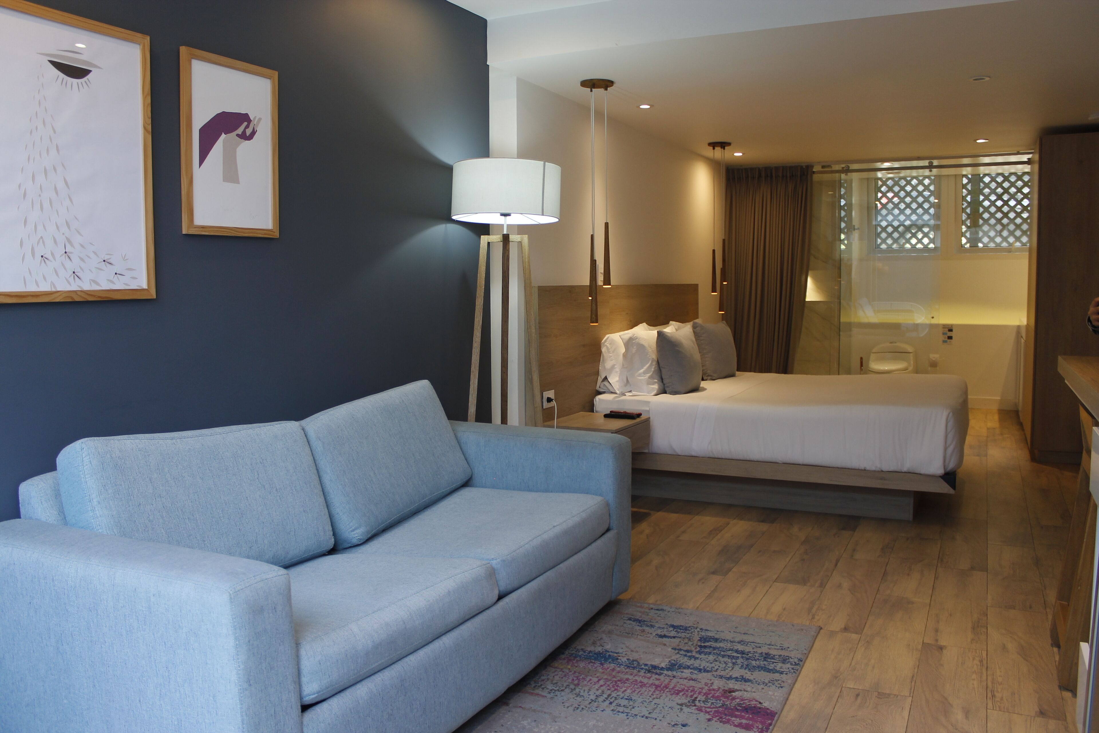Una habitación de hotel moderna con un sofá azul claro, una cama con ropa de cama blanca, una lámpara de pie y obras de arte en la pared.