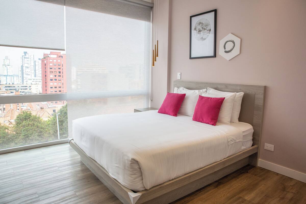 Ein modernes Schlafzimmer mit großem Fenster, einem Bett mit weißer und rosa Bettwäsche und Blick auf die Stadt.