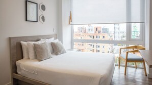 A modern bedroom with a large bed, a wooden chair, and a city view through the window.