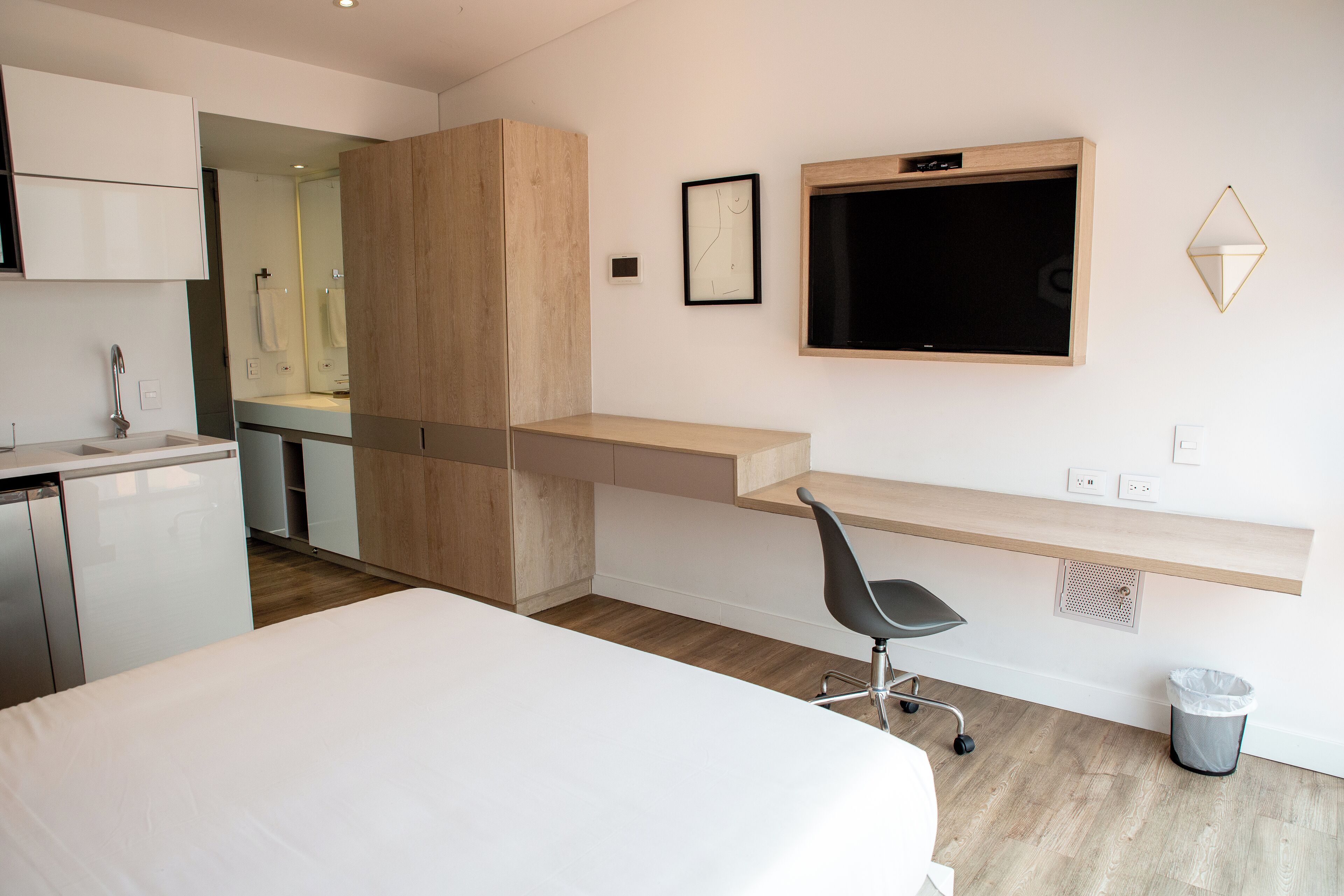 A modern hotel room with a bed, a desk with a chair, a TV, and a small trash bin.