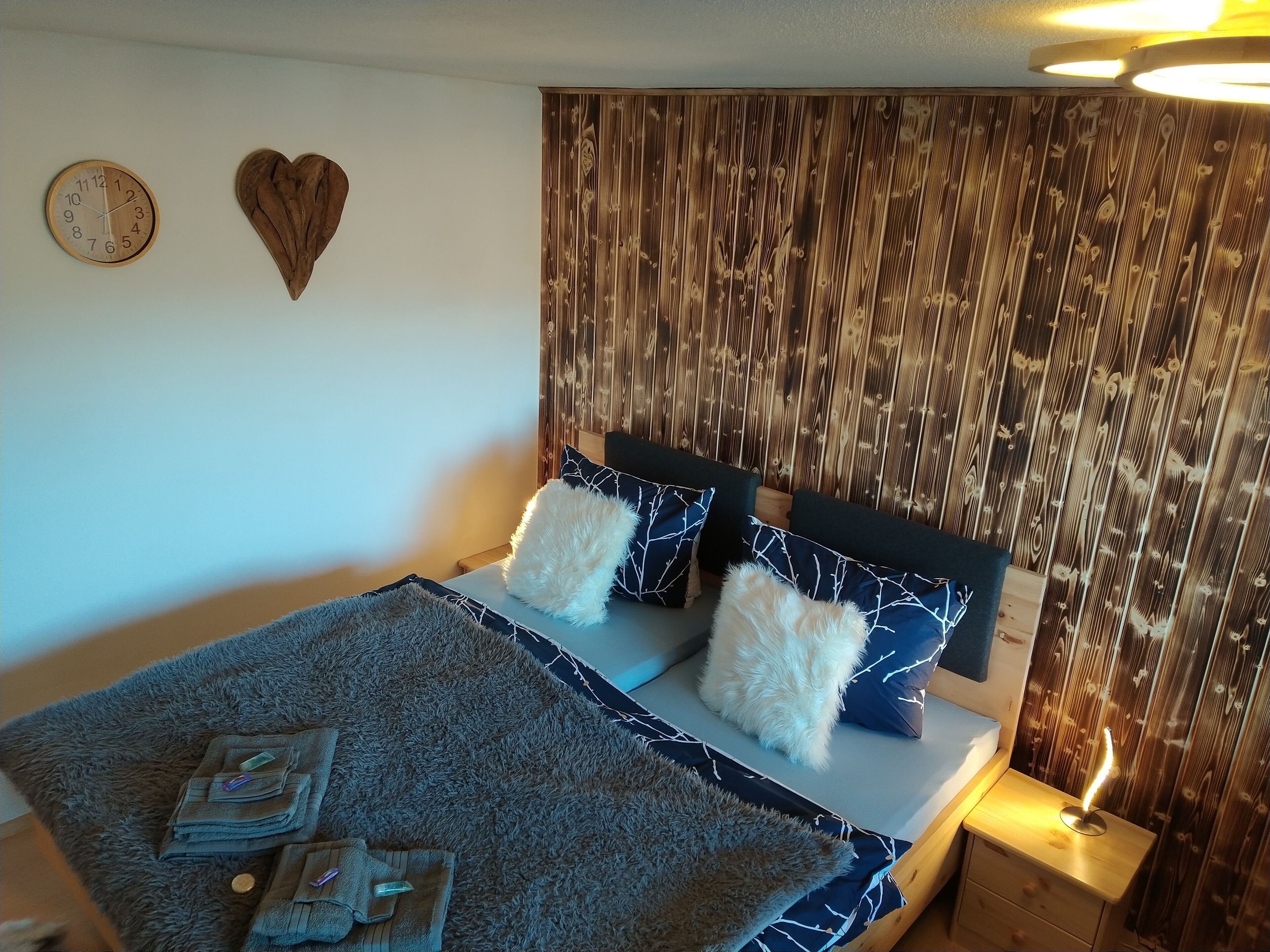 Ein ordentlich bezogenes Bett mit Holz Kopfteil, einem flauschigen weißen Kissen und einem blauen Kissen mit Muster. Ein kleiner Nachttisch mit Lampe ist sichtbar.