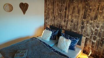 Ein ordentlich bezogenes Bett mit Holz Kopfteil, einem flauschigen weißen Kissen und einem blauen Kissen mit Muster. Ein kleiner Nachttisch mit Lampe ist sichtbar.