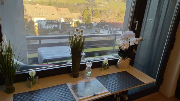 Una habitación con un gran ventanal que da a un paisaje montañoso, una pequeña mesa con un tapete, un jarrón con flores blancas y un cuadro enmarcado.
