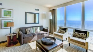 A modern hotel room with a large window offering a sea view, a sofa set, a side table with a lamp, and framed artwork on the wall. - Two Bedroom Luxury Suite, Ocean View, Virginia Beach, Virginia (2555337) (Virginia Beach)