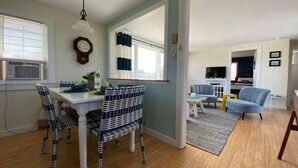 Ein Essbereich mit einem weißen Tisch und gemusterten Stühlen, einer Uhr an der Wand und Blick in ein Wohnzimmer mit einem blauen Sofa und einem Fernseher.