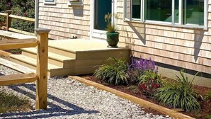 Ein eingeschossiges Haus mit Veranda, einem kleinen Garten mit verschiedenen Pflanzen und einem Holzzaun.
