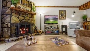 Un salon chaleureux avec une cheminée en pierre, un téléviseur à écran plat fixé au mur, un fauteuil confortable et une table basse agrémentée d’objets décoratifs.