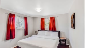 Une chambre à coucher avec un lit, deux fenêtres ornées de rideaux rouges, une table de chevet et une œuvre d’art accrochée au mur.