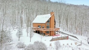 A wooden cabin with a large deck and multiple windows, surrounded by a snowy forest. - Hot Tub & Sauna ~ The Ridge Chalet ~ Wood Fireplace ~ Sleeps 12 (Great Valley)