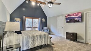 A bedroom with a sloped ceiling, a bed with a patterned bedspread, a nightstand, a chair, a dresser, a TV on the wall, and a window with a view of trees. - Hot Tub & Sauna ~ The Ridge Chalet ~ Wood Fireplace ~ Sleeps 12 (Great Valley)