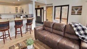 A modern living room with a brown leather sofa, wooden coffee table, and a kitchen area with white cabinets and a bar stool. - Hot Tub & Sauna ~ The Ridge Chalet ~ Wood Fireplace ~ Sleeps 12 (Great Valley)
