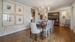 Ein Esszimmer mit einem Holztisch, gedeckt für vier Personen, umgeben von weißen Stühlen. Über dem Tisch hängt eine Chandelüster, und zur Rechten befindet sich ein Bücherregal mit Büchern und Dekoration.