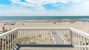 Vue sur une plage depuis une terrasse en bois dotée d’une balustrade, avec une plage de sable et l’océan en arrière-plan.