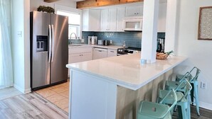 Une cuisine moderne avec des armoires blanches, un réfrigérateur en acier inoxydable, un plafond en bois et des chaises de couleur turquoise.