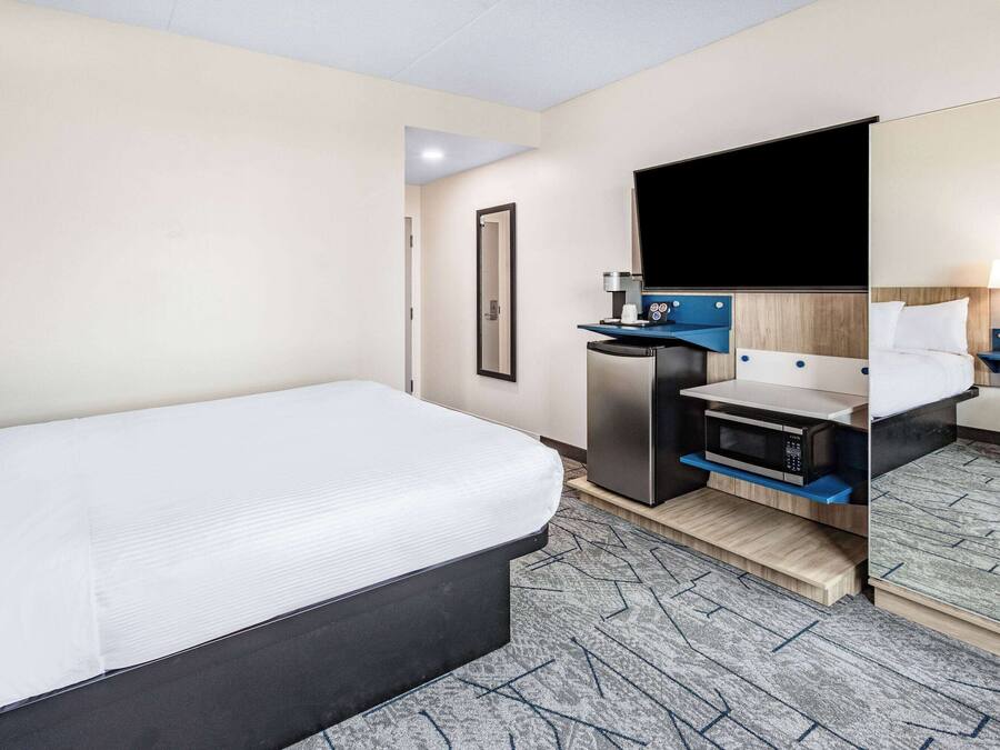 A hotel room with a large bed, a desk with a microwave, a mirror, and a nightstand with a lamp.