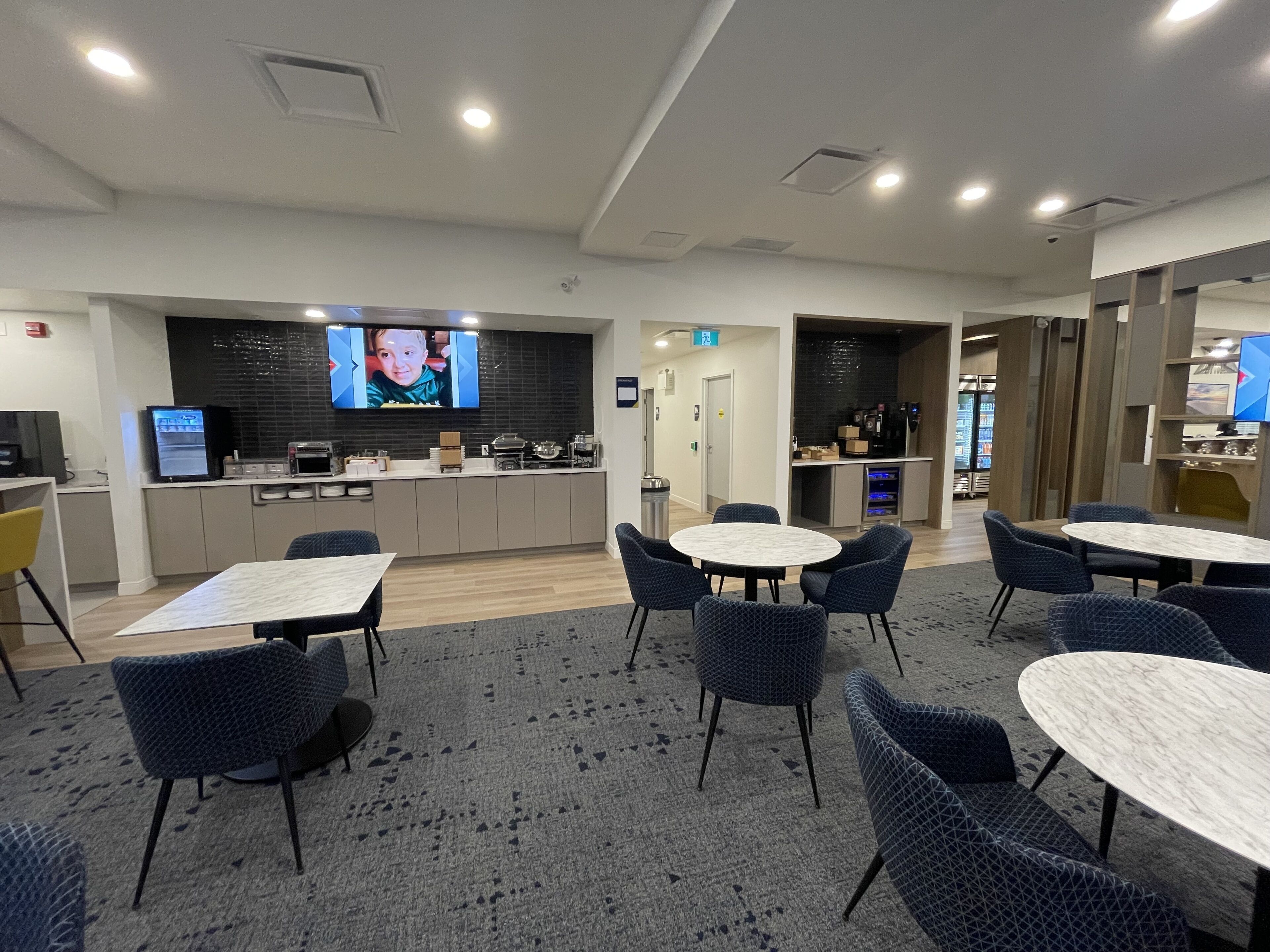 A modern hotel lobby with a kitchen area, seating arrangements, and a television screen.