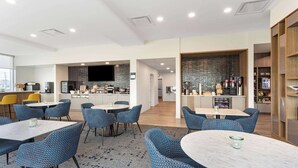 Un moderno hall de hotel con mesas redondas y sillas de diseño azul, una zona de cocina con televisor de pantalla plana y una estación de café con diversas bebidas.