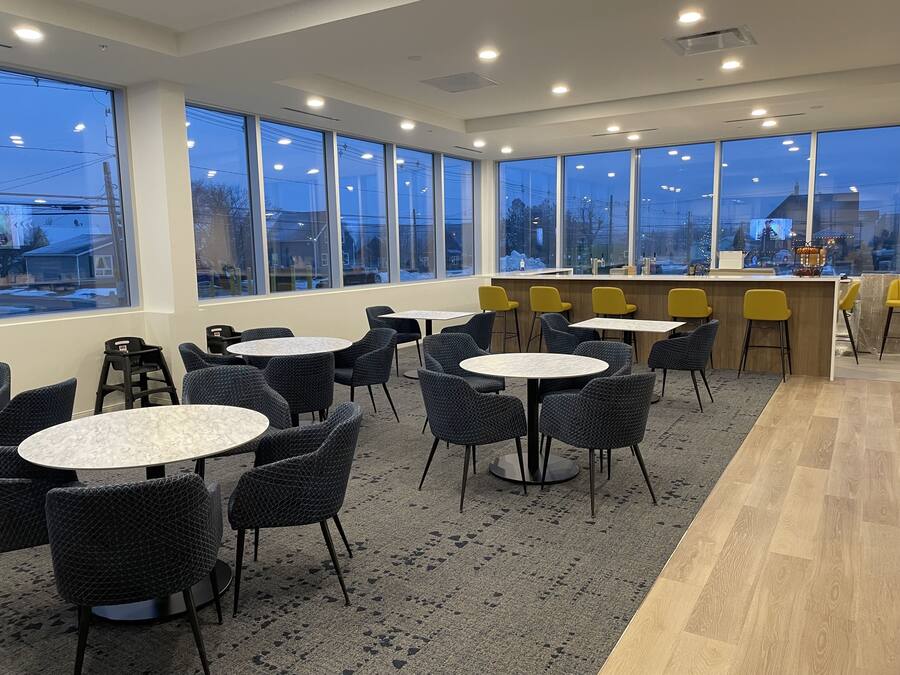 Un espace de détente intérieur moderne, avec des tables et des chaises rondes, de grandes fenêtres offrant une vue sur l’extérieur et un comptoir de bar avec des chaises jaunes.