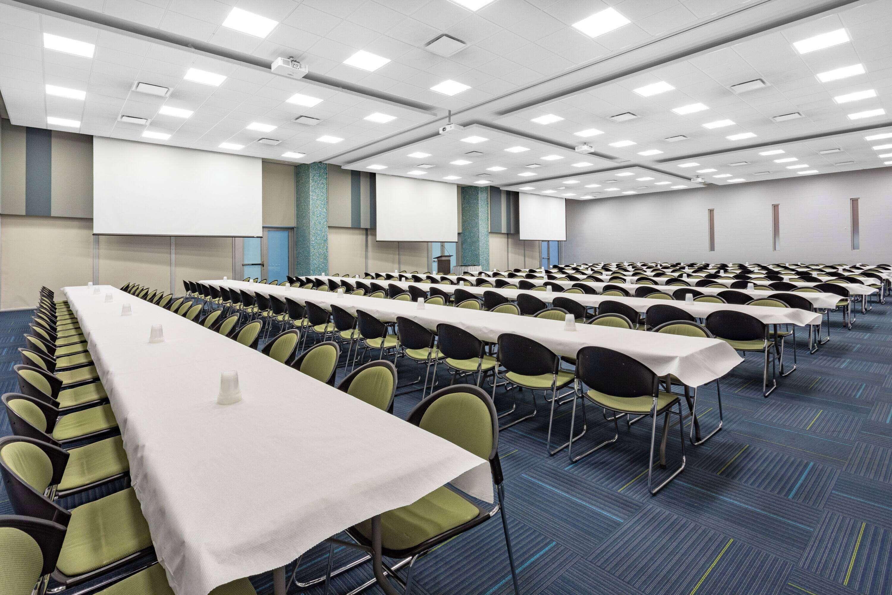 Een congreszaal met rijen tafels en stoelen, een scherm voor een beamer en een vloerbedekking.
