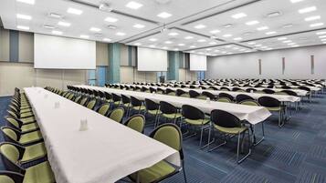 Een congreszaal met rijen tafels en stoelen, een scherm voor een beamer en een vloerbedekking.