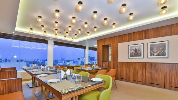 Un espace repas moderne doté de plusieurs tables et chaises, de grandes fenêtres offrant une vue sur la ville et d’un mur lambrissé en bois orné de tableaux encadrés.