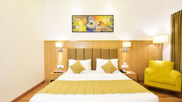 A hotel room with a large bed, a wooden headboard, two bedside tables with lamps, a yellow armchair, and a colorful abstract painting on the wall.