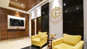 Un hall d’entrée avec deux fauteuils jaunes, une table ronde ornée d’un vase décoratif, un téléviseur fixé au mur et une grande horloge murale.