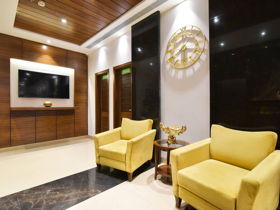 A lobby with two yellow armchairs, a round table with a decorative bowl, a mounted TV, and a large wall clock.
