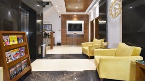 Un hall d’entrée moderne avec un plafond en bois, un grand téléviseur à écran plat fixé au mur, un coin salon avec des canapés jaunes et un présentoir à magazines contenant diverses publications.