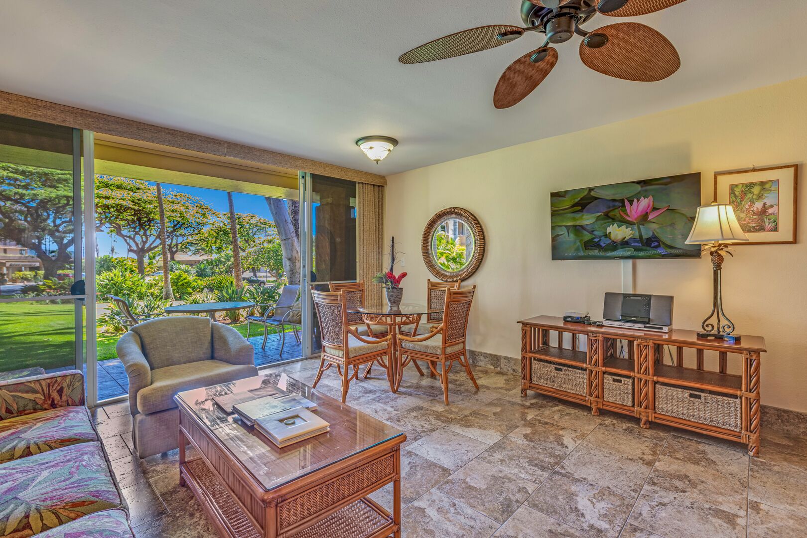 A spacious living area with a glass door leading to a garden, a ceiling fan, a dining table with chairs, a sofa, a coffee table, a TV, and a lamp.