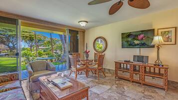 A spacious living area with a glass door leading to a garden, a ceiling fan, a dining table with chairs, a sofa, a coffee table, a TV, and a lamp.