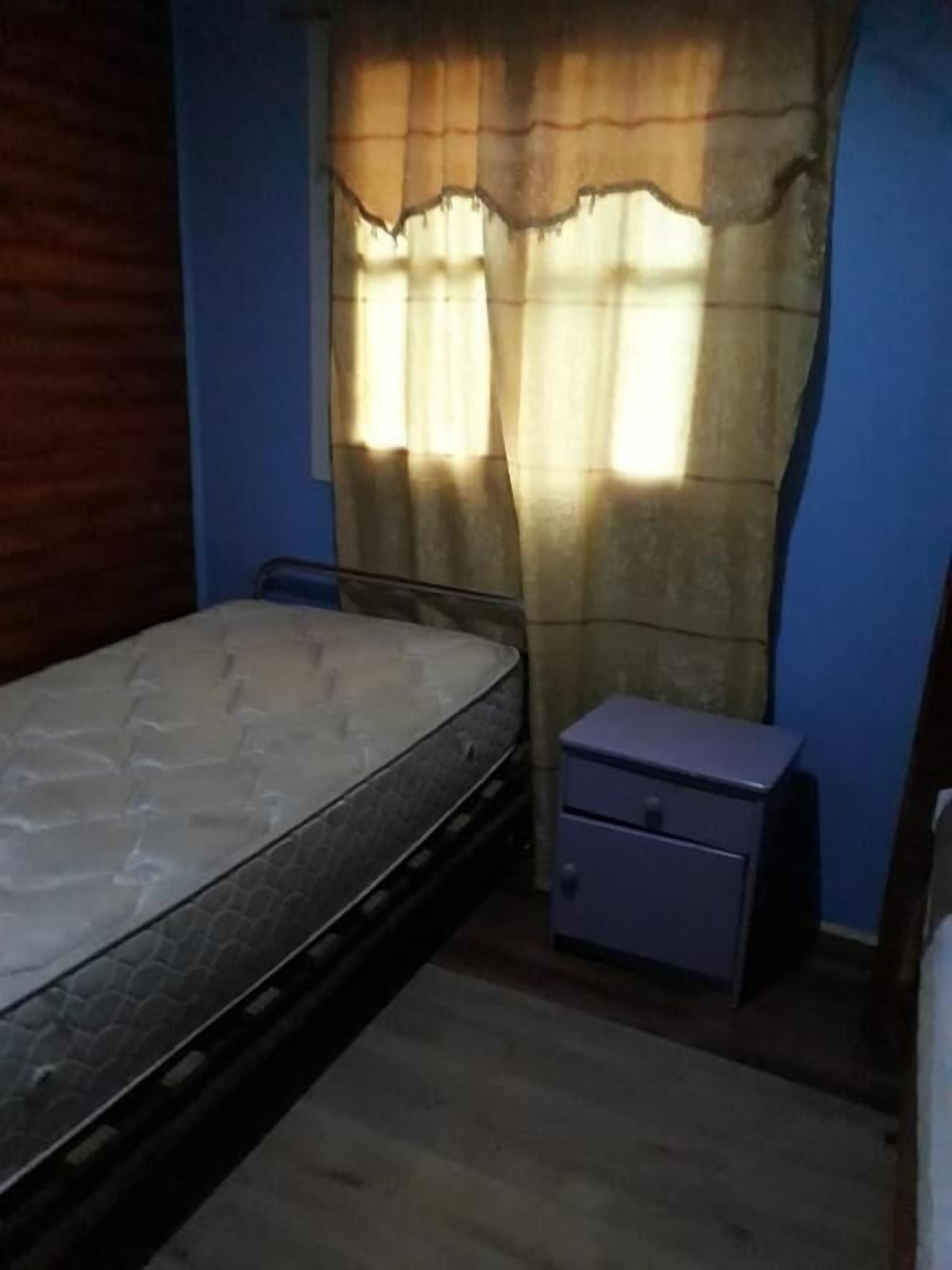 A small, dimly lit room with a single bed, a nightstand, and a window with curtains.