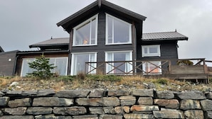 Une maison moderne à deux étages, dotée de grandes fenêtres, d’un mur en pierre au premier plan et d’une terrasse en bois.