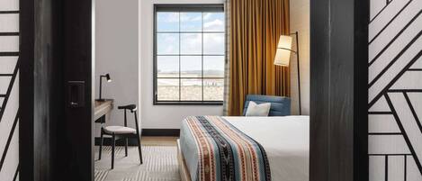 A hotel room with a large window, a bed with a patterned blanket, a desk with a chair, and a lamp.