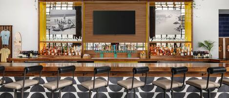 A bar area with a large screen, bar stools, and a variety of bottles.