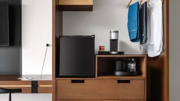 A modern hotel room with a black refrigerator, a coffee maker, and a shelf with water bottles. There is a wooden desk with a chair and a flat-screen TV mounted on the wall. Clothes are hanging on a rack.