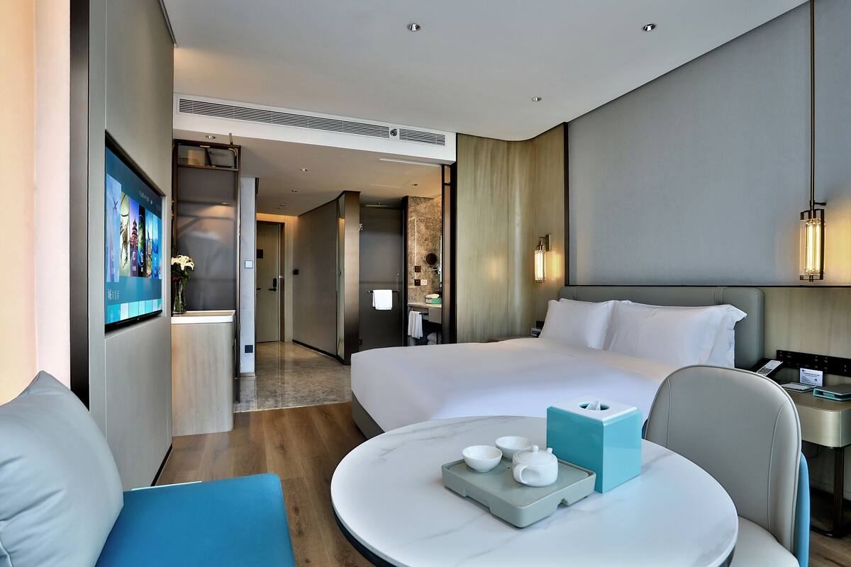 Una habitació d'hotel moderna amb un llit gran, un escriptori amb un ordinador, una taula rodona amb una bandeja de tasses de te, i un televisor fixat a la paret.