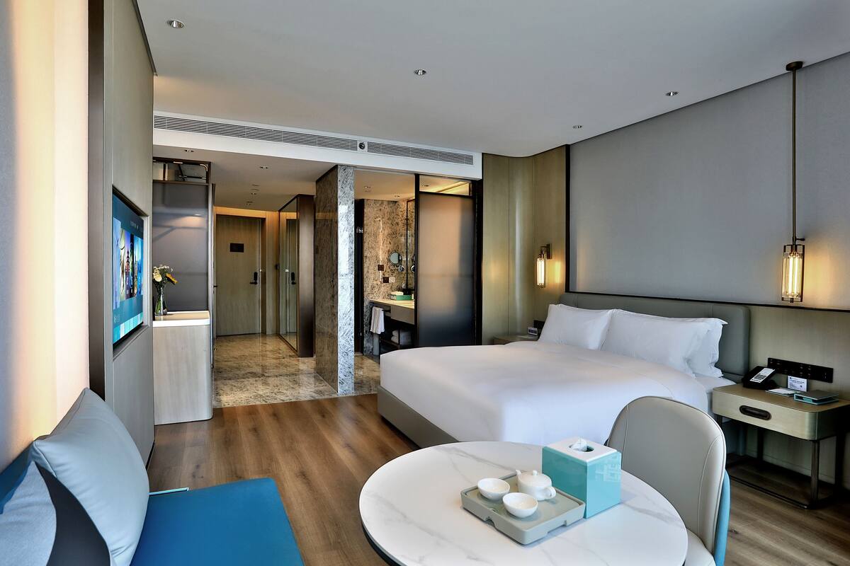 Habitació d'hotel moderna amb un llit gran, una taula de nit amb una làmpada, un escriptori amb un ordinador i vista al passadís.