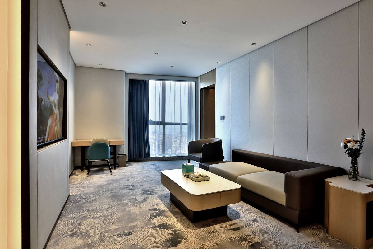 Habitació d'hotel moderna amb sofà, taula de cafè, escriptori i una finestra gran amb cortines.