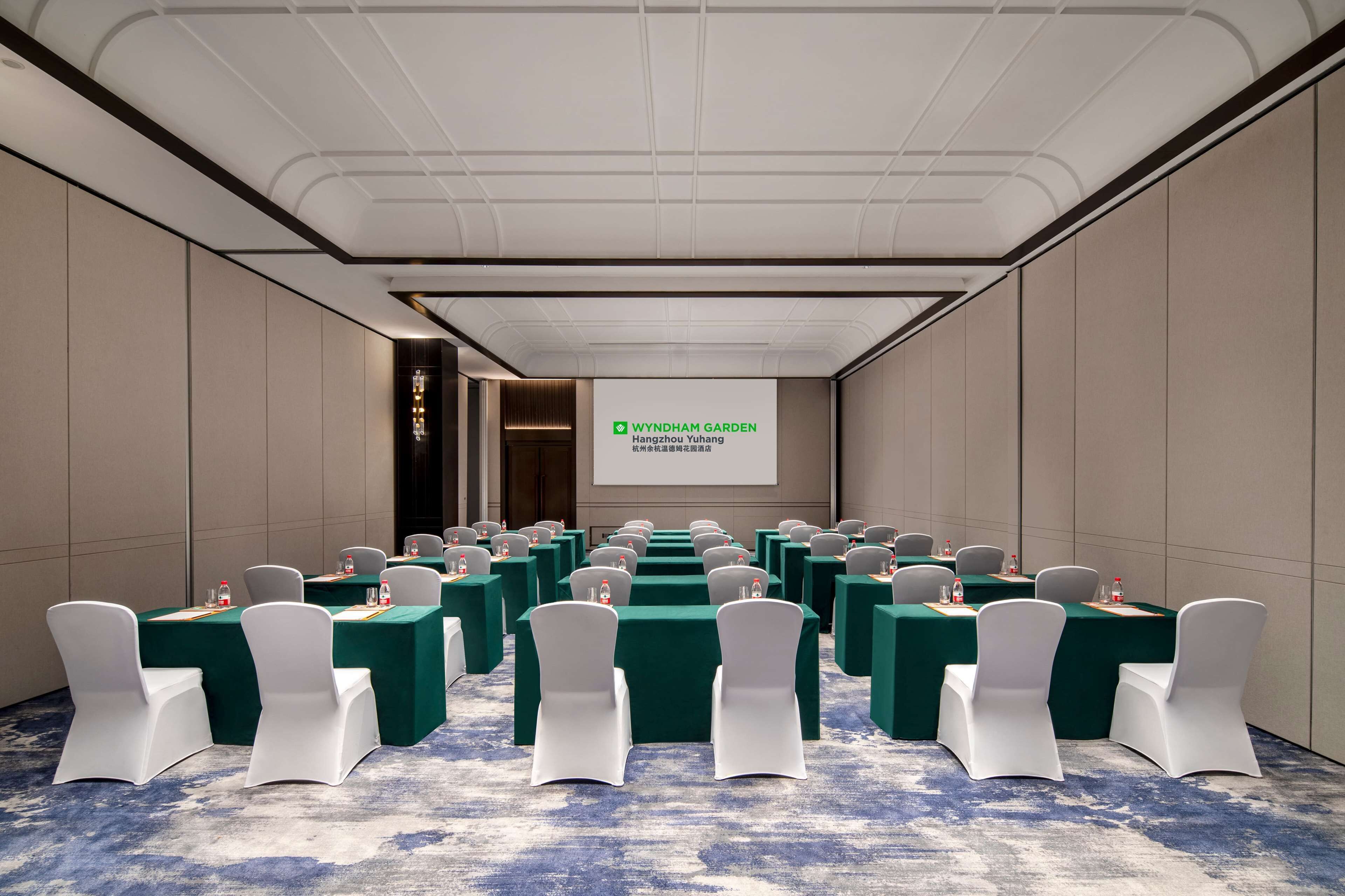 Un salón de conferencias con filas de mesas y sillas, una pantalla de proyector y suelo alfombrado.