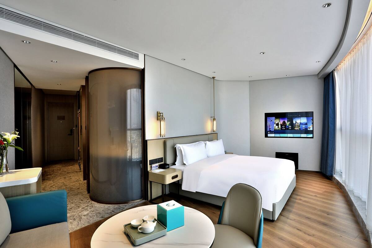 Una habitació d'hotel moderna amb un llit gran, una televisió de pantalla plana, una taula de treball amb una cadira i vista a la ciutat per la finestra.