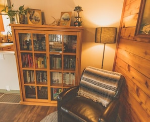 A cozy room with a leather chair, a wooden bookshelf filled with books, a lamp, and a framed picture on the wall.
