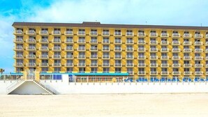 A large, multi-story hotel with a uniform yellow facade, balconies on each floor, and a covered walkway with blue chairs. - Two Bedroom Ocean Front Condo, Ormond Beach 2555514 (Ormond Beach)