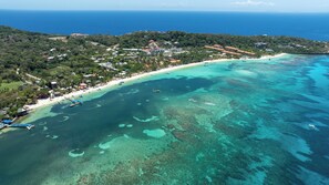 An aerial view of a coastal area with a beach, clear turquoise waters, and a forested hill. - Aqua de Mare #1 steps away from West Bay Beach (West Bay)