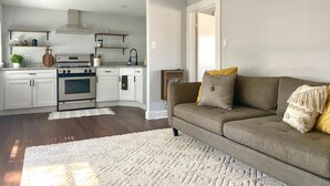 Una cocina moderna con armarios blancos, un horno empotrado y una estufa. Hay una tabla de cortar de madera y una planta en maceta sobre la encimera. Un sofá color beige con cojines amarillos y grises se encuentra en la zona de estar.