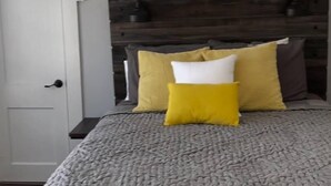 Un dormitorio con una cama, cabecera de madera, colcha gris acolchada y unidad de aire acondicionado blanca sobre la cama.