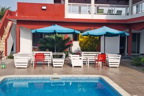 A swimming pool area with white and red lounge chairs, blue umbrellas, and a staircase leading to a balcony. - JADE - ASSINIE (Assinie)