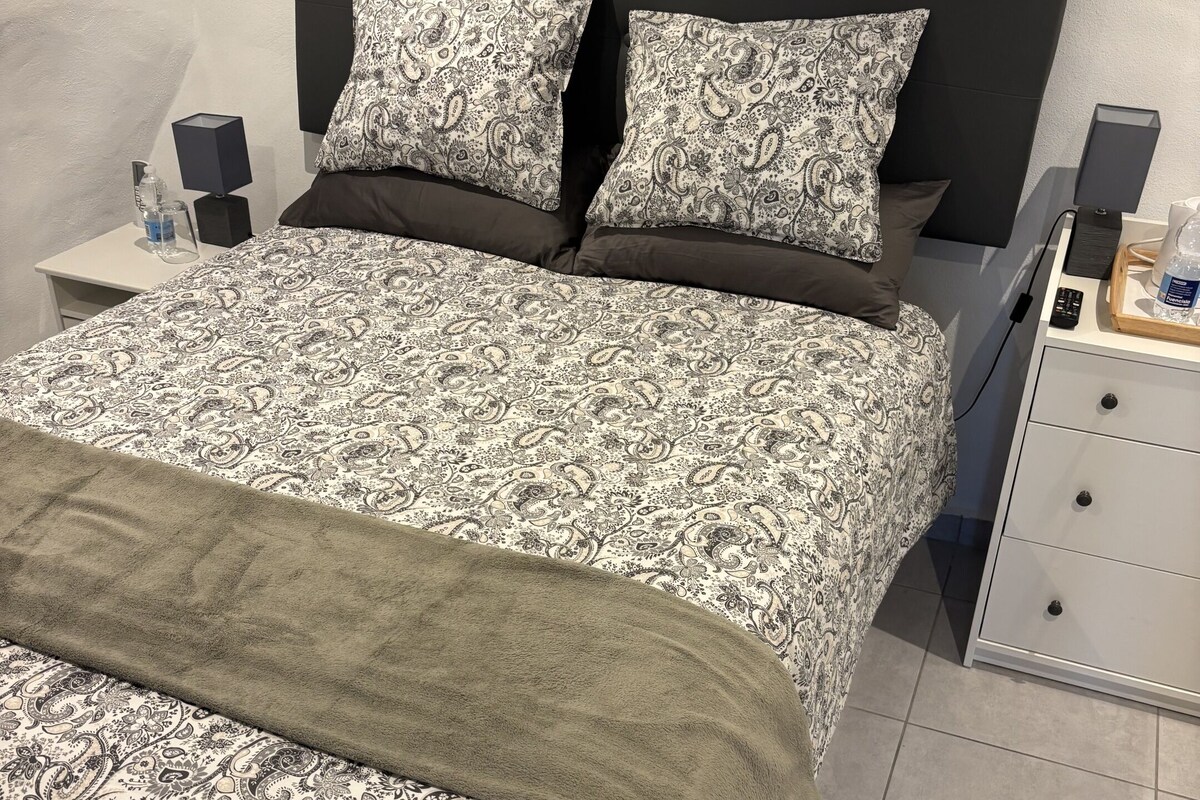 Un lit bien fait, avec une couvre-lit à motifs et des oreillers, une table de chevet avec une lampe, et une petite commode placée à côté du lit.